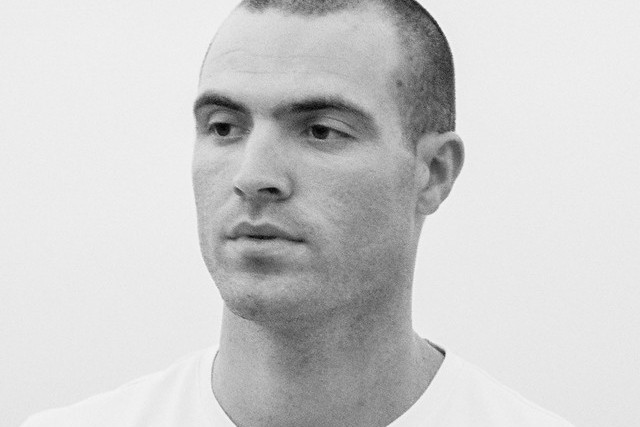 Emmit Fenn in a black-and-white portrait, close-cropped hair and a white t-shirt, looking off to his left against a plain pale wall