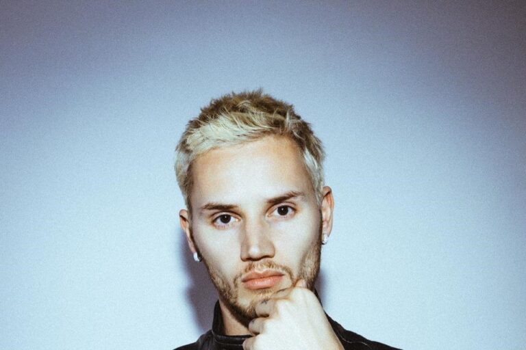 Mau P press portrait, bleached blonde hair and a black leather vest over a white tee, resting his fist against his chin against a pale blue-grey wall