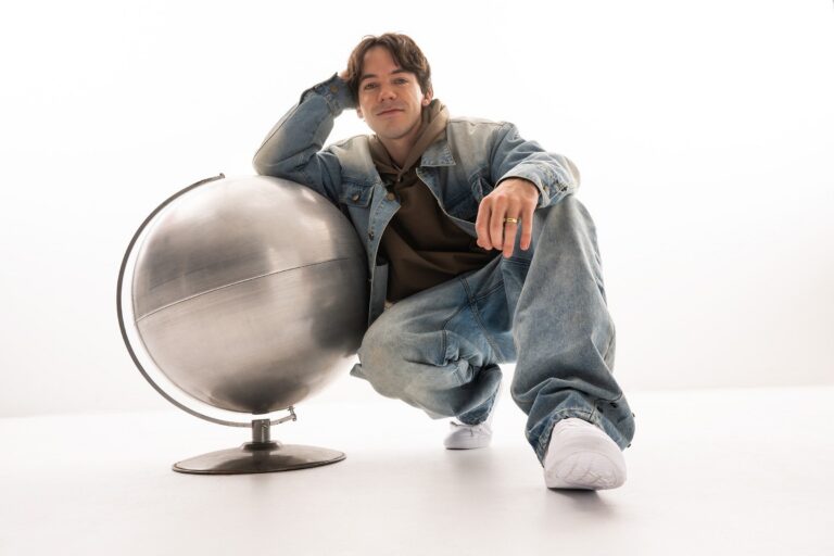 Connor Price crouching beside a large brushed-metal desk globe against a plain white studio backdrop, wearing an oversized light-wash denim jacket and baggy jeans over an olive hoodie, with white sneakers