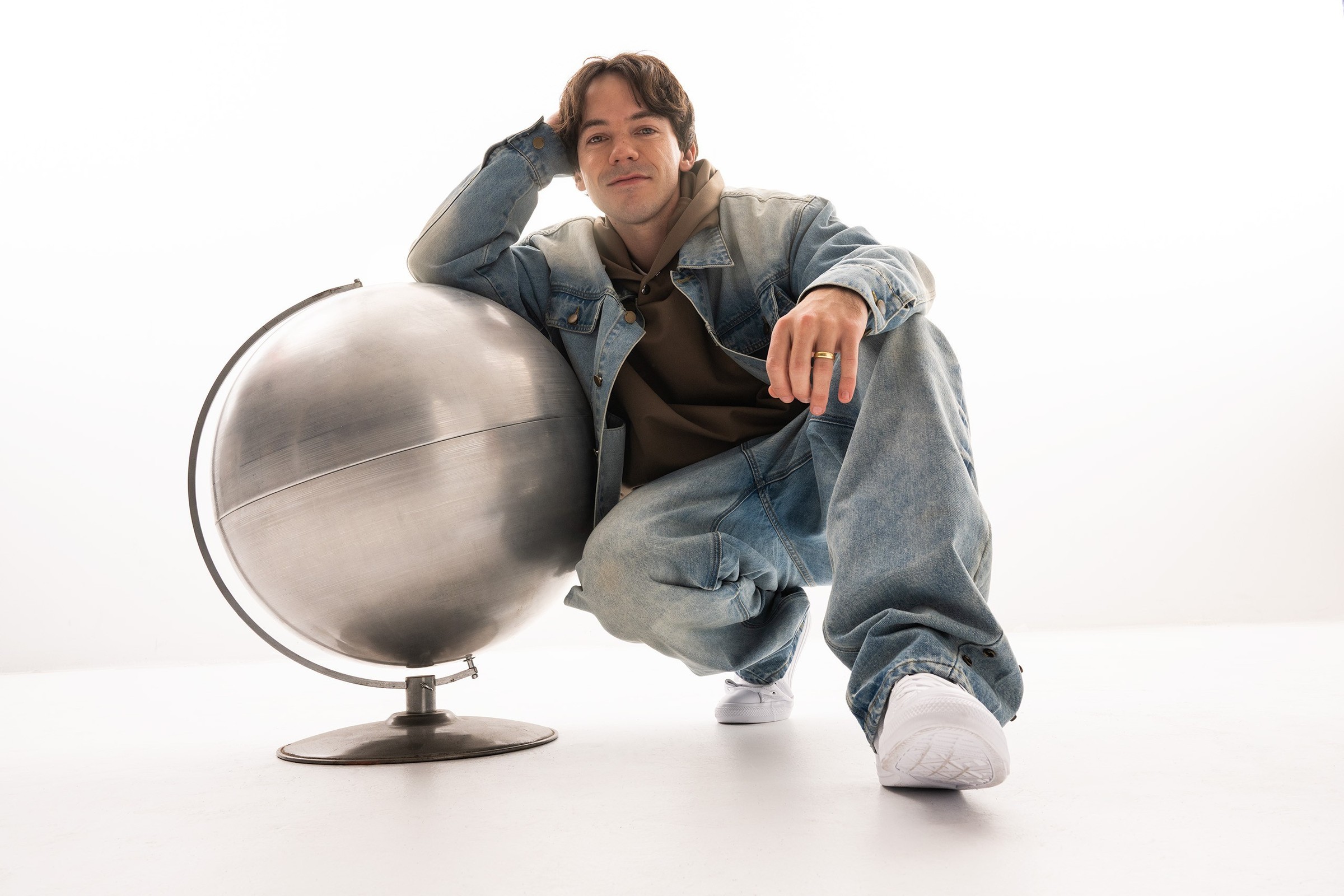 Connor Price crouching beside a large brushed-metal desk globe against a plain white studio backdrop, wearing an oversized light-wash denim jacket and baggy jeans over an olive hoodie, with white sneakers