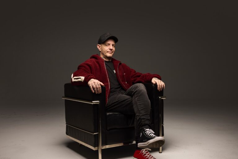 deadmau5 seated in a black leather and chrome armchair in a grey studio, wearing a black star-print cap, dark red fleece jacket over a black tee, black jeans, and mismatched black and red Converse high-tops
