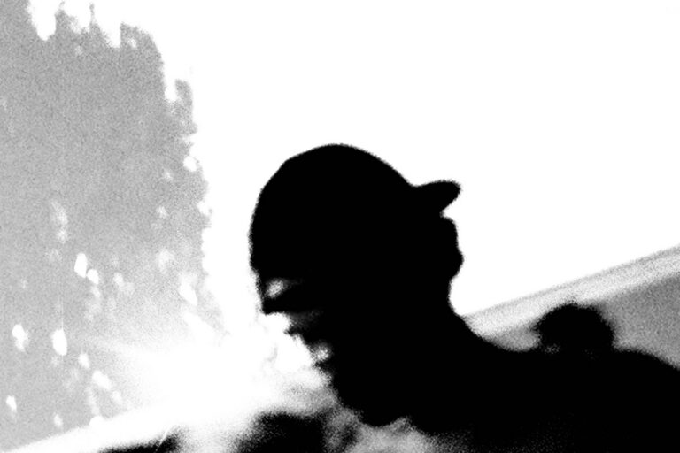 Arden Jones in high-contrast black and white, shown in profile in a baseball cap against a blown-out white sky, with a blurred tree line at left