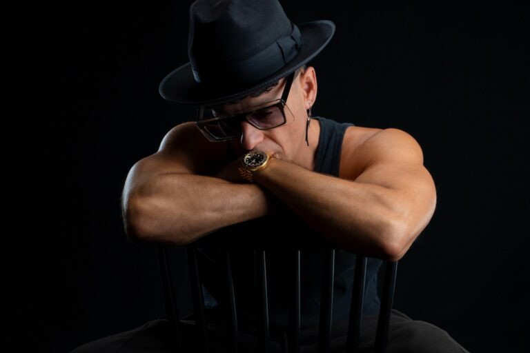 Timmy Trumpet in a black fedora and tinted glasses, arms folded over the back of a black chair against a black backdrop