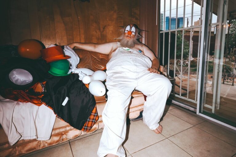 Cover art for Wiz Khalifa single Yiccen, a man in white clown face paint and an orange nose reclining in white overalls on a worn couch on a patio with party hats and balloons piled beside him
