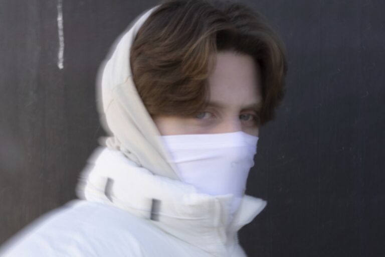 Aaron Hibell press portrait in a white hooded puffer jacket and white face covering, motion-blurred against a dark wall