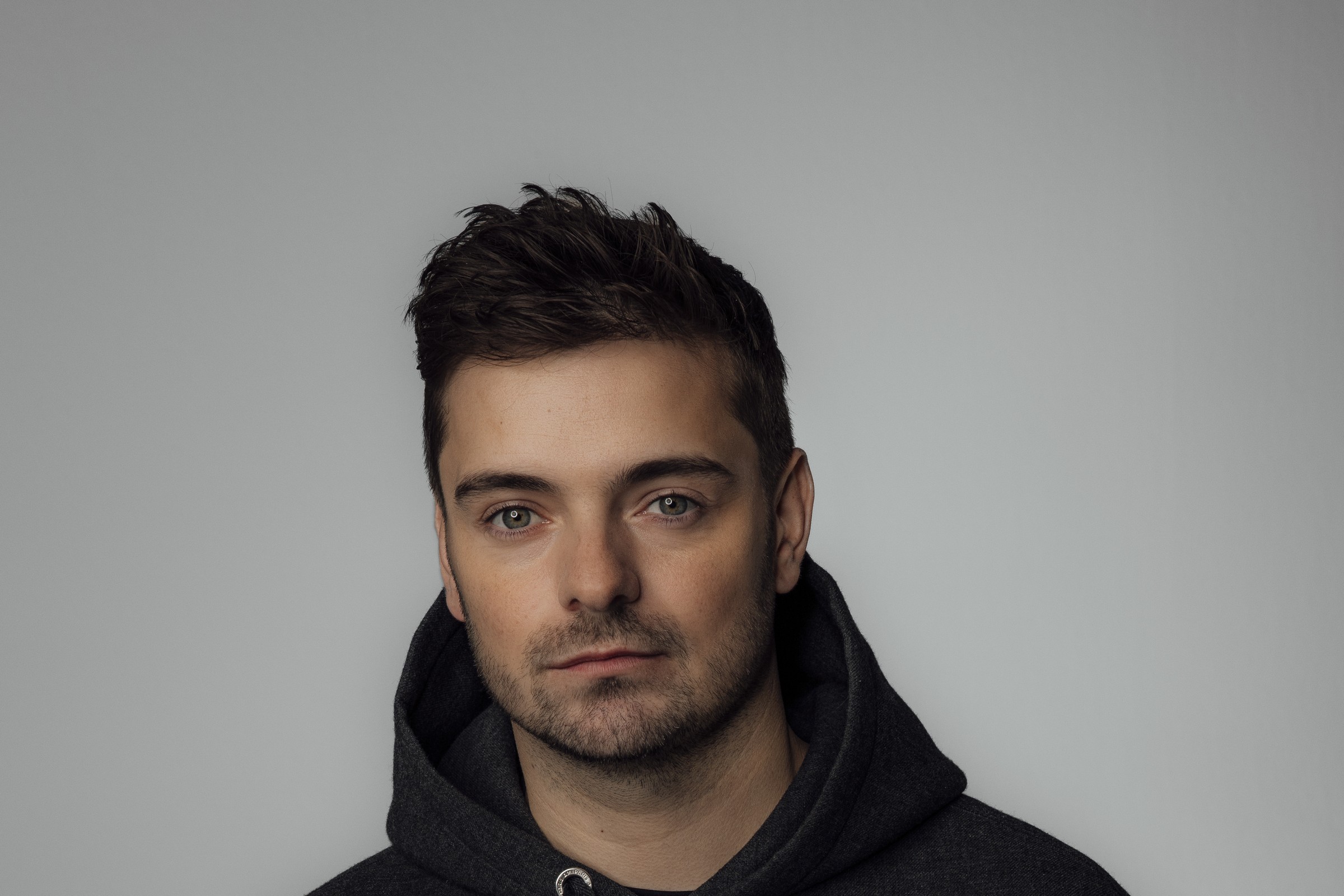 Martin Garrix press portrait in a charcoal hooded sweatshirt, looking at the camera against a plain light-grey studio backdrop