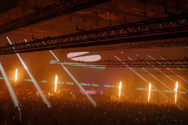 A packed crowd under amber light at an A State of Trance show, with flame jets firing along the stage, white beams cutting through haze, and 'A State of Trance' text repeated across the stage screens beneath the ASOT logo