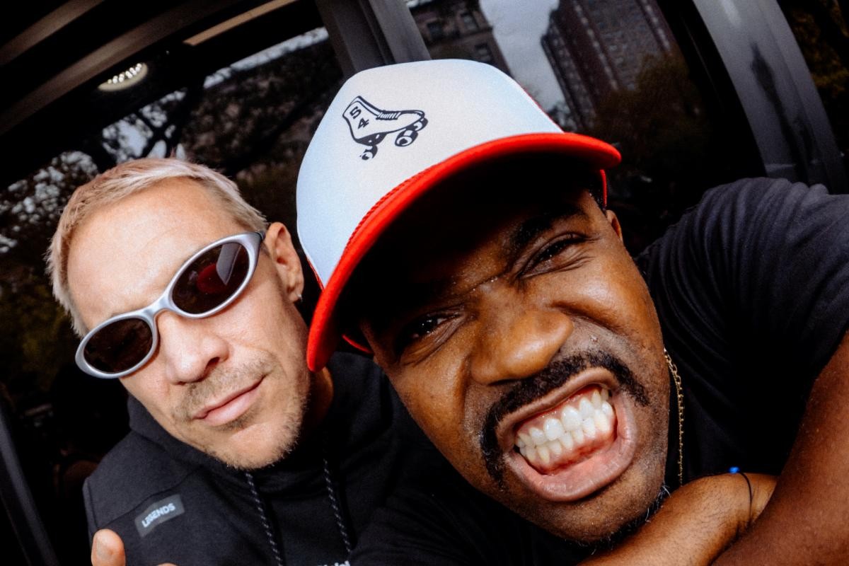 Diplo in silver wraparound sunglasses and a black hoodie leans in beside a grinning A$AP Ferg in a white and red trucker cap, with city buildings behind them