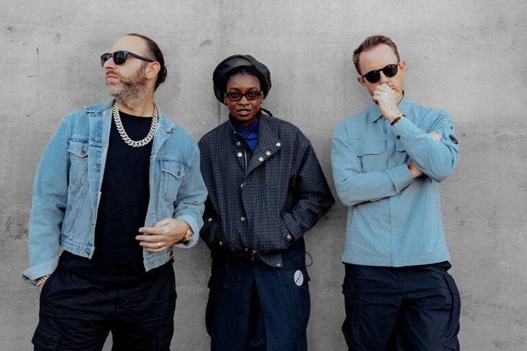 Chase & Status and Little Simz stand against a grey concrete wall: Saul Milton in sunglasses, a denim jacket and a chunky chain on the left, Little Simz in a black beret, glasses and a dark check jacket in the centre, Will Kennard in sunglasses and a light blue overshirt with a hand to his chin on the right