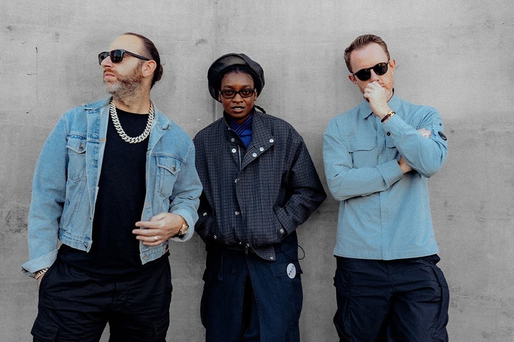 Chase & Status and Little Simz stand against a grey concrete wall: Saul Milton in sunglasses, a denim jacket and a chunky chain on the left, Little Simz in a black beret, glasses and a dark check jacket in the centre, Will Kennard in sunglasses and a light blue overshirt with a hand to his chin on the right