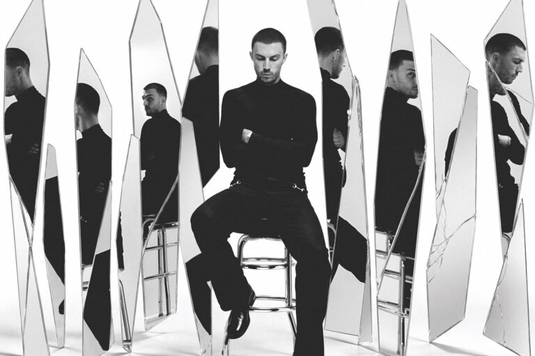 Cloonee in a black turtleneck seated on a metal stool, arms crossed, surrounded by tall shattered mirror shards reflecting him from different angles, black and white