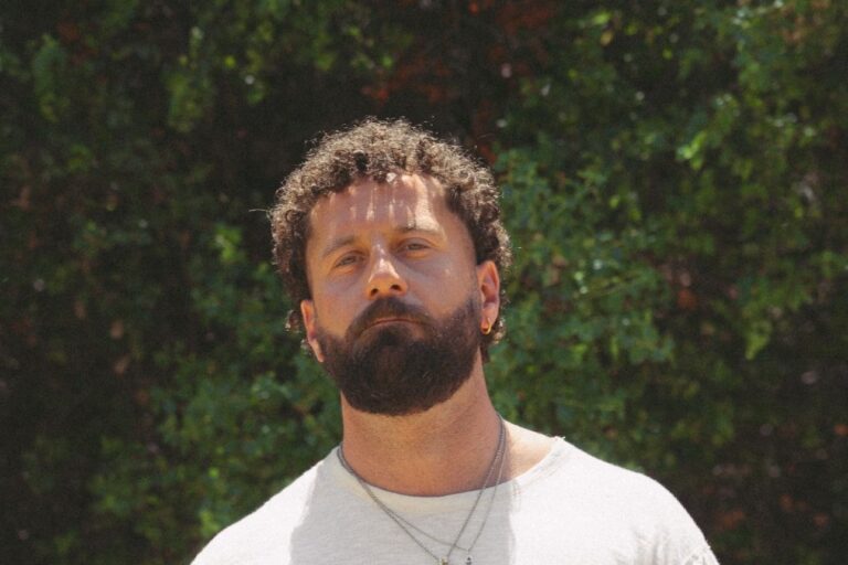 Elderbrook press portrait, curly hair and full beard, in a white T-shirt and silver chain necklaces in front of a sunlit green hedge