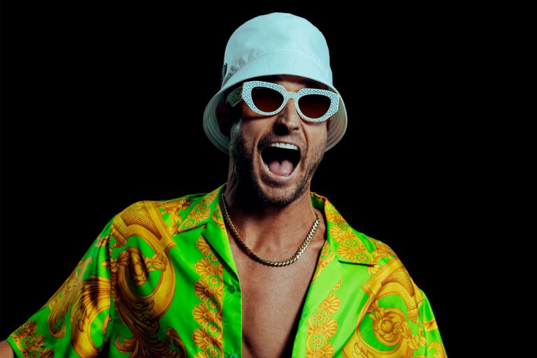 FISHER press portrait, mouth wide open mid-shout, in a white bucket hat, white jewelled cat-eye sunglasses, gold chain and an open neon-green and gold baroque silk shirt against a black backdrop