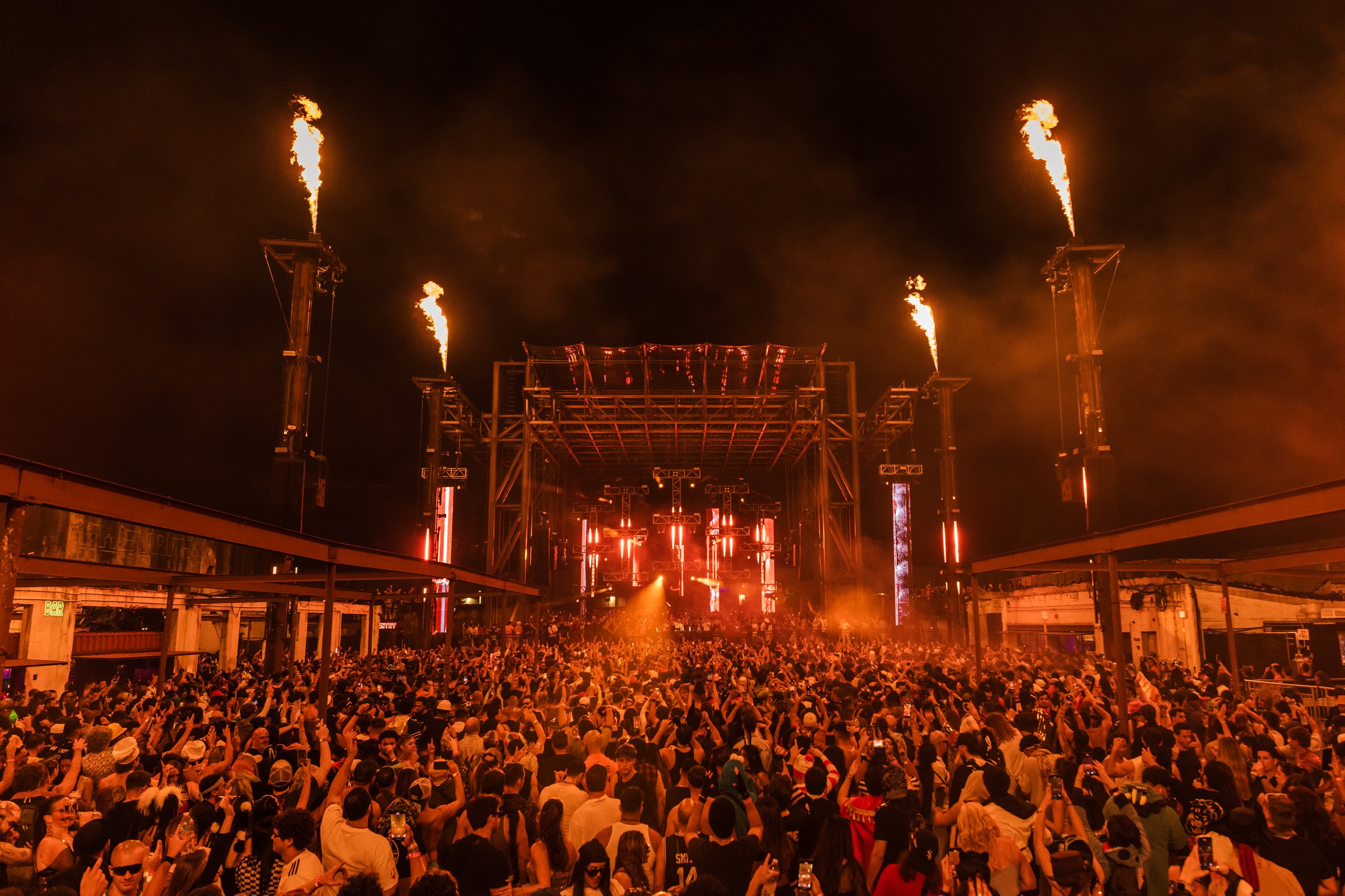Packed crowd in Halloween costumes facing the Infinity Room's steel-truss stage at Factory Town, with flame cannons firing from four towers and red light haze