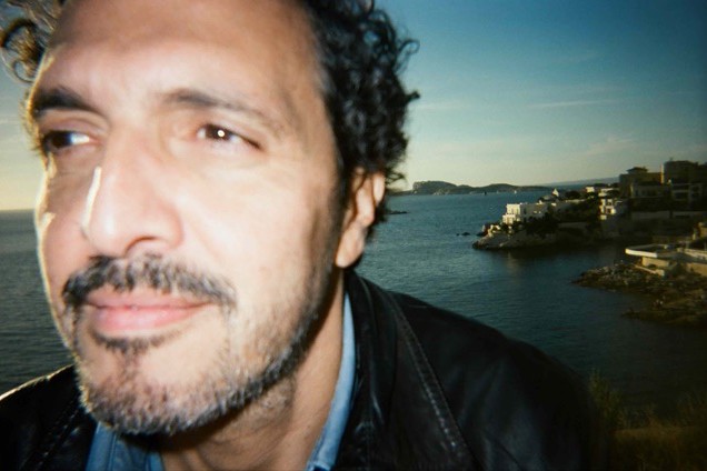 Kid Francescoli in a close-up film-style portrait, curly dark hair and a stubbled beard, in a black leather jacket, with the sea and a rocky coastline at sunset behind him