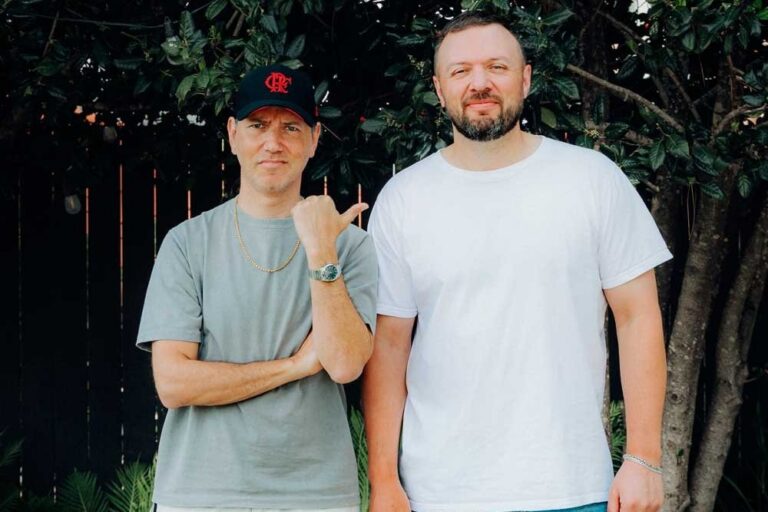 Tiga and Chris Lake standing side by side in front of dark leafy trees and a wooden fence. Tiga, on the left, wears a black cap with a red logo, a grey T-shirt and a gold chain, and points his thumb toward Lake. Lake, on the right, has a beard and wears a white T-shirt.