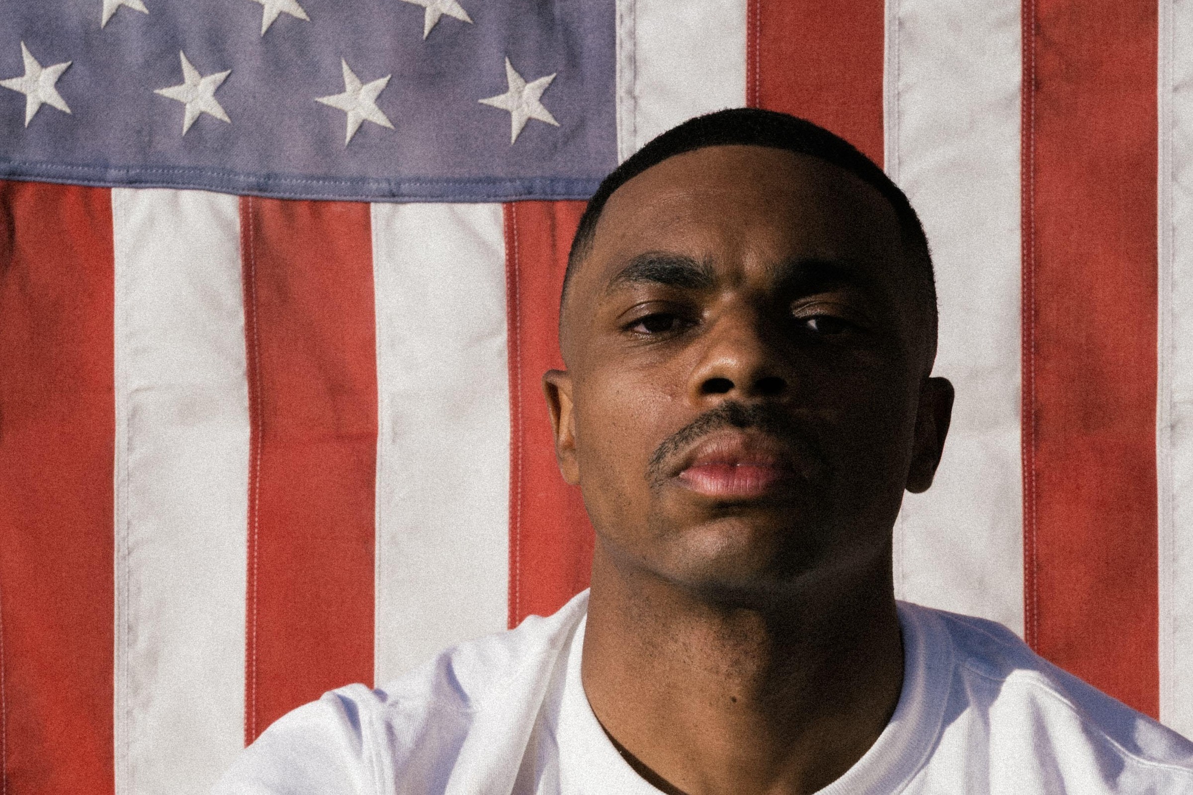 Vince Staples press portrait in a white T-shirt, looking down into the camera in front of a hanging American flag