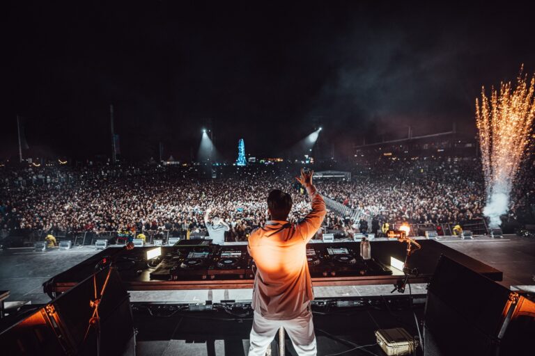 Kygo seen from behind at the DJ booth with one arm raised, facing a huge nighttime festival crowd, with sparkler pyrotechnics on the right and spotlights cutting through haze