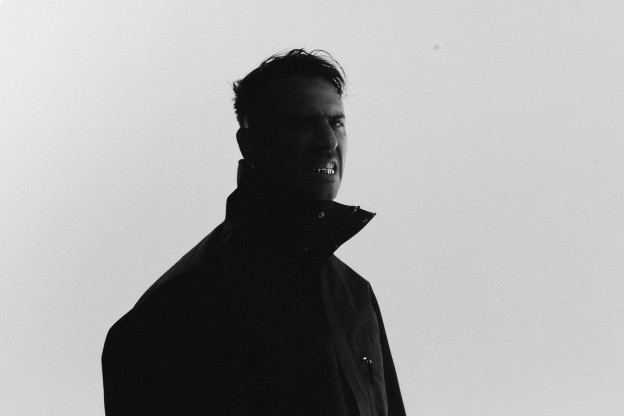 Boys Noize black and white press portrait in a dark high-collared jacket, silhouetted against a pale grey sky