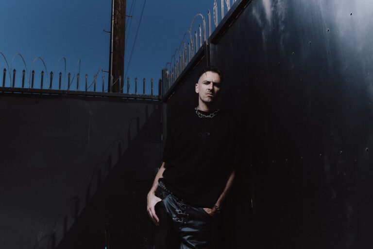 Boys Noize press portrait in a black T-shirt, chain necklace and buckled leather trousers, leaning against a black metal wall beneath a razor-wire fence and a deep blue sky