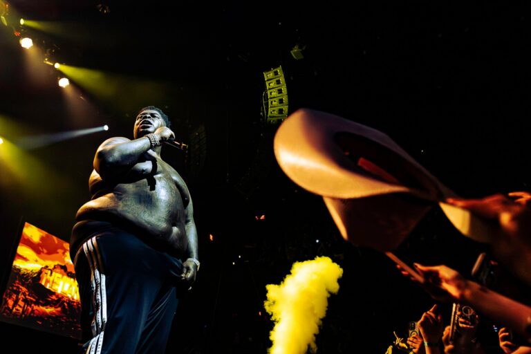 BigXthaPlug performing shirtless onstage under yellow stage lights, microphone raised, as a fan in the foreground waves a tan cowboy hat beside a plume of yellow smoke