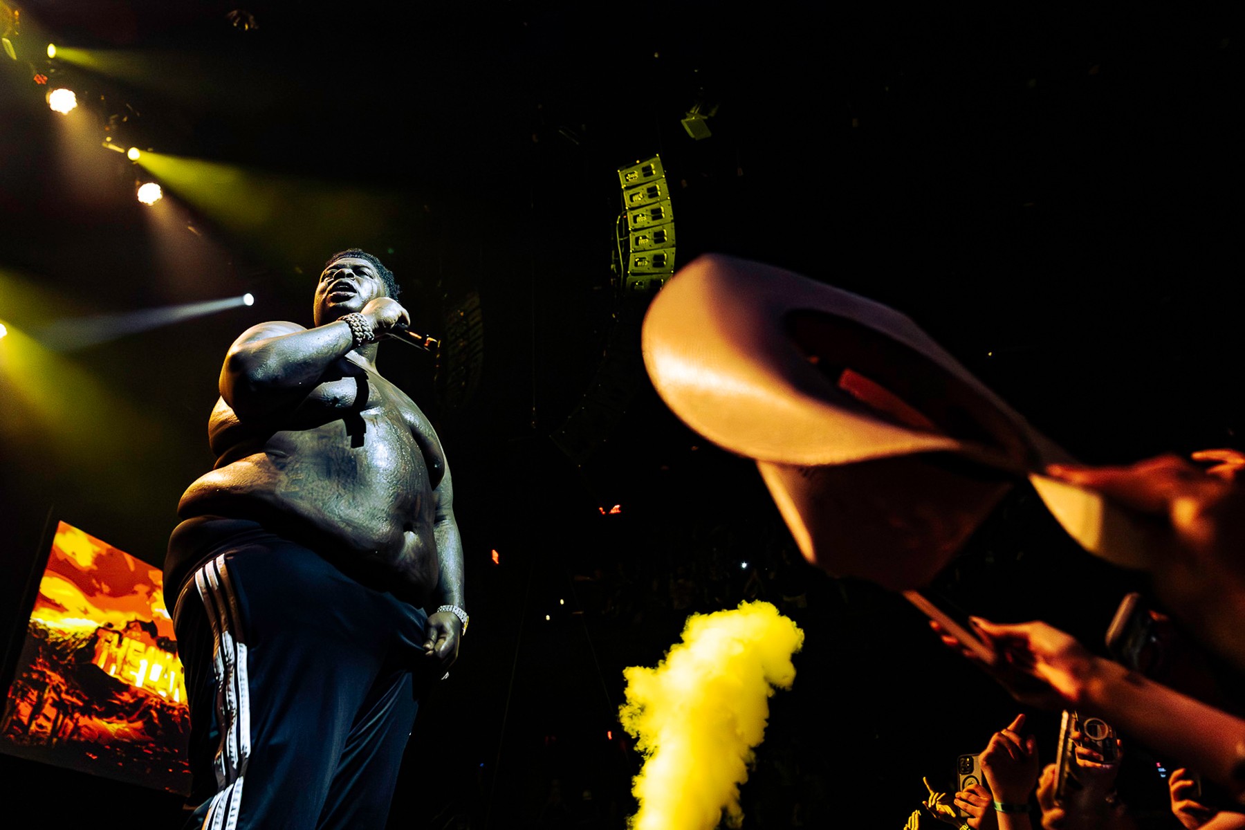 BigXthaPlug performing shirtless onstage under yellow stage lights, microphone raised, as a fan in the foreground waves a tan cowboy hat beside a plume of yellow smoke