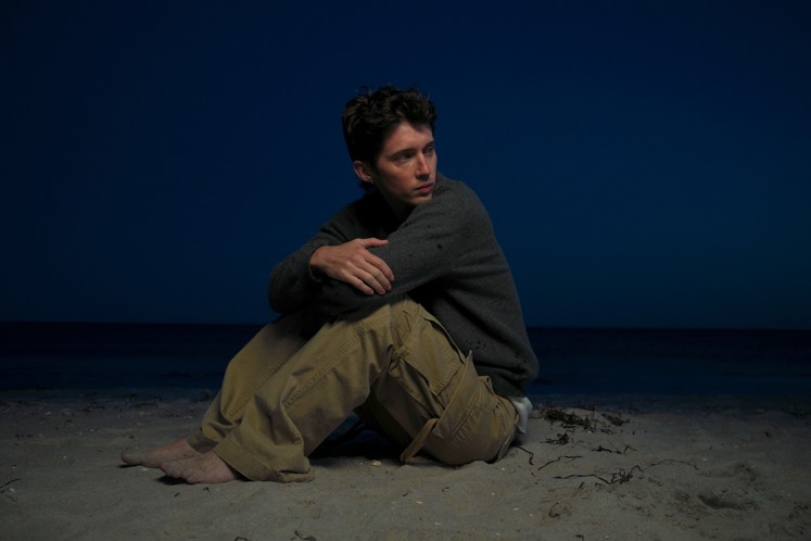 Troye Sivan sitting barefoot on a beach at dusk in a dark grey sweater and khaki cargo pants, arms wrapped around his knees, looking off to the side against a deep blue sky and sea