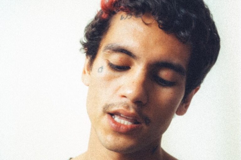 Dominic Fike press portrait, eyes cast down and lips parted, curly dark hair with a streak of red, against a pale washed-out backdrop
