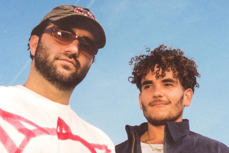ANOTR duo press photo shot from below against a blue sky: one member in a brown cap, amber sunglasses and a white tee with red print, the other with curly hair in a navy zip jacket and gold chain