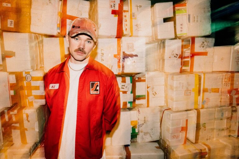Dom Dolla press portrait in a worn red trucker cap and red Sea-Land work jacket over a white T-shirt, standing in front of stacked white foam shipping boxes taped in orange and red