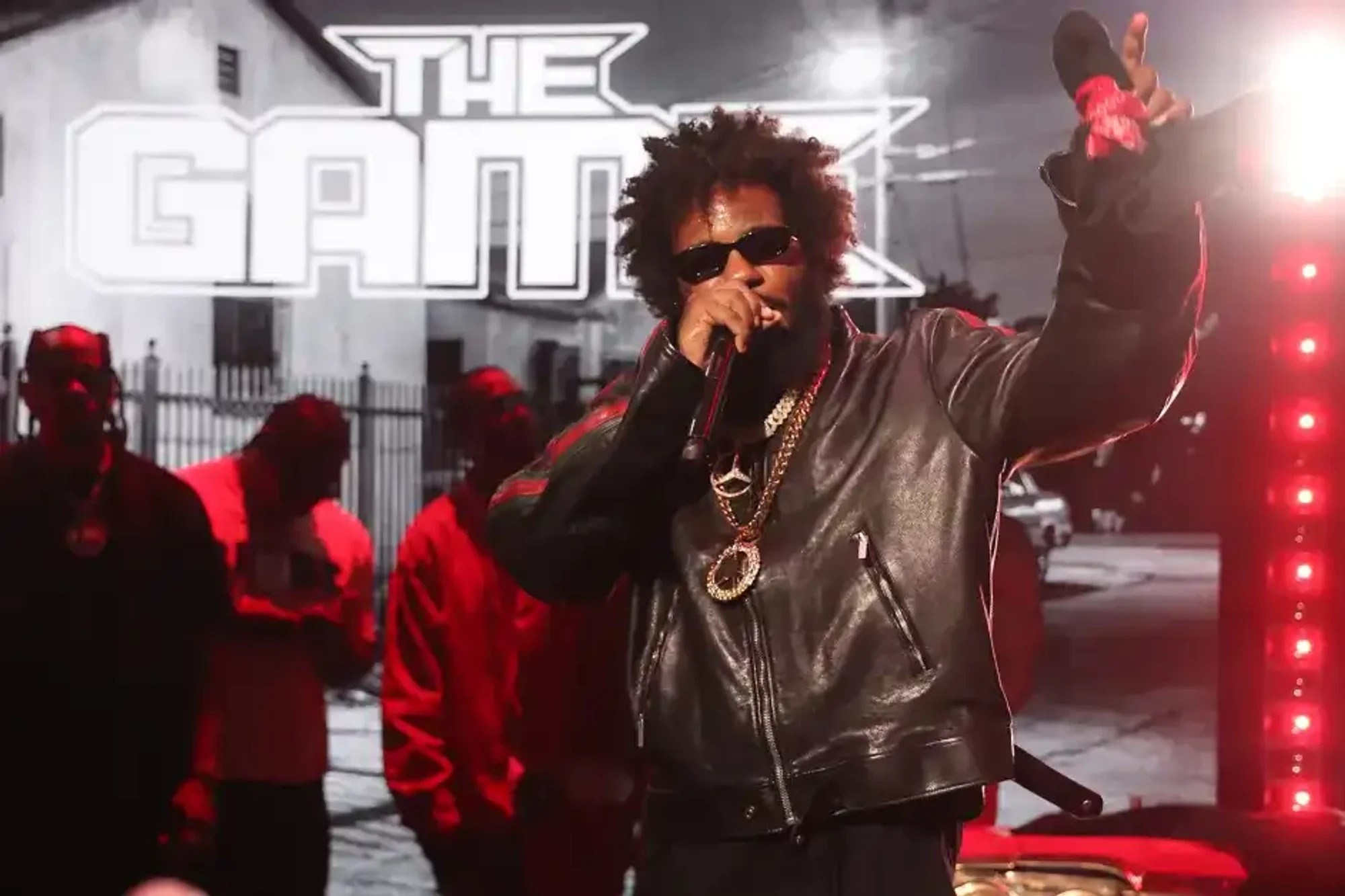 The Game performing on stage in sunglasses and a black leather jacket, mic in hand and arm raised, a giant 'THE GAME' logo over a black-and-white Compton street scene behind him and red-lit crew at left