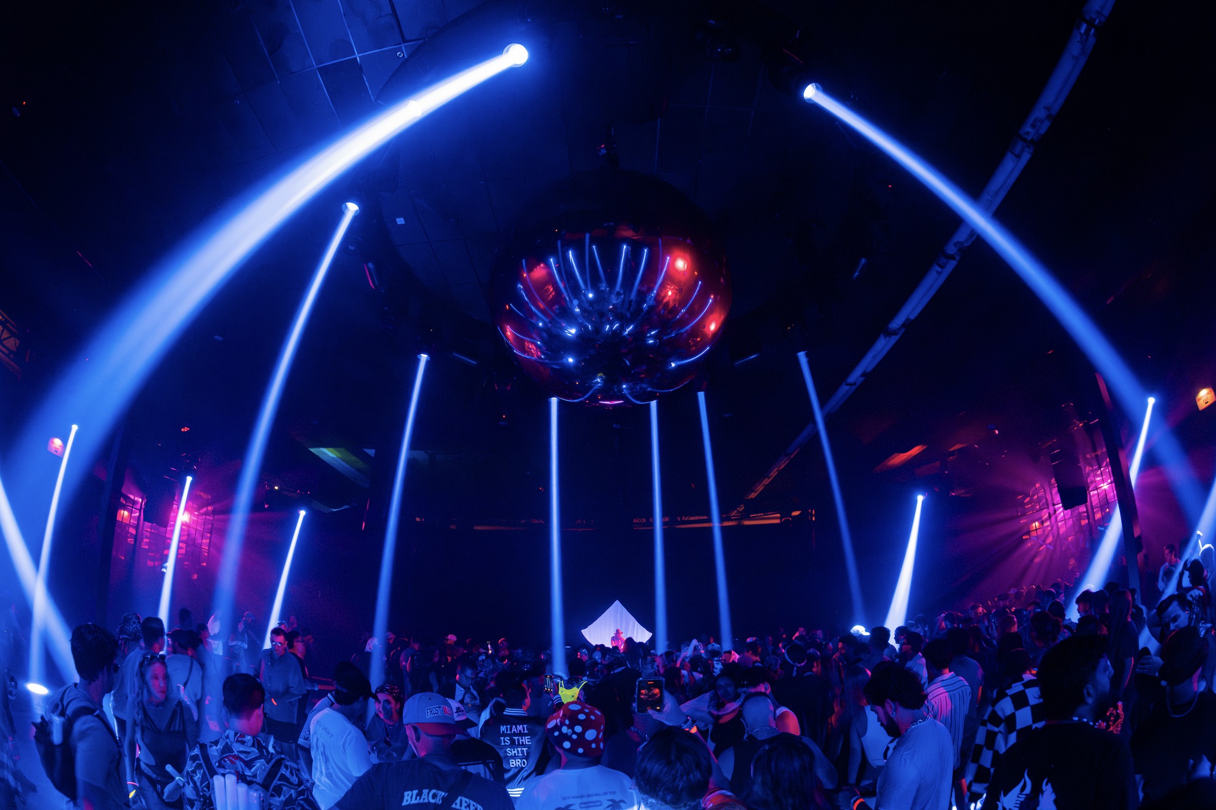 Crowd under blue light beams and a large mirrored sculpture hanging from the ceiling inside the III Points ::444:: warehouse stage
