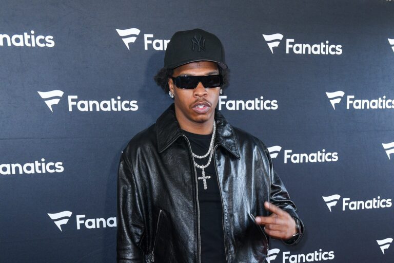 Lil Baby in a black NY cap, black wraparound sunglasses, a black leather jacket and a silver cross chain, posing in front of a Fanatics step-and-repeat backdrop