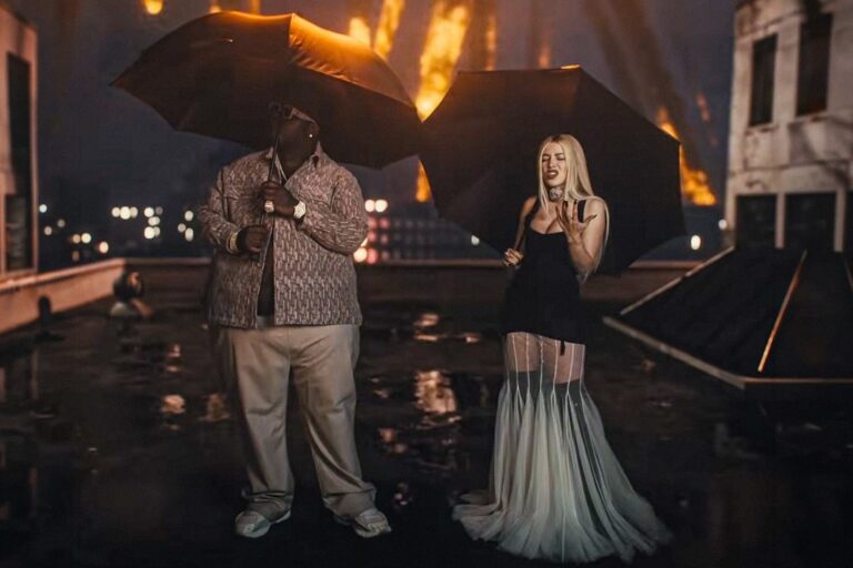 BigXthaPlug and Ava Max under black umbrellas on a rain-soaked rooftop at night, flames burning in the background, from the BURN music video