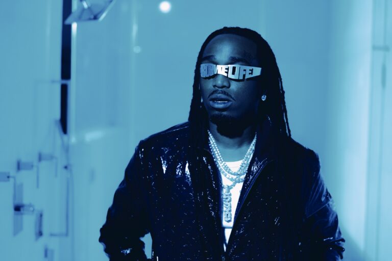 Quavo press photo in a blue-tinted hallway, wearing silver cut-out QRÖMELIFE letter sunglasses, a glossy black leather jacket and stacked diamond chains