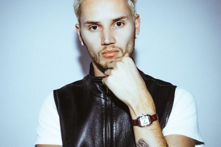 Mau P press portrait with bleached blond hair, a black leather zip vest over a white T-shirt, his hand at his chin, against a pale blue-grey wall
