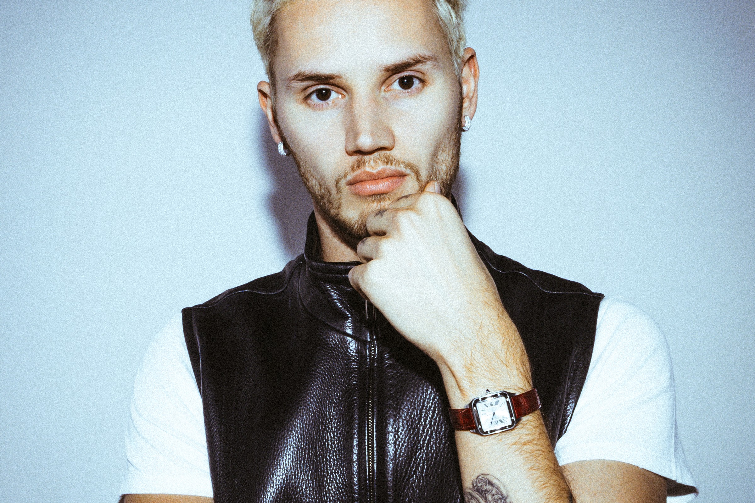 Mau P press portrait with bleached blond hair, a black leather zip vest over a white T-shirt, his hand at his chin, against a pale blue-grey wall