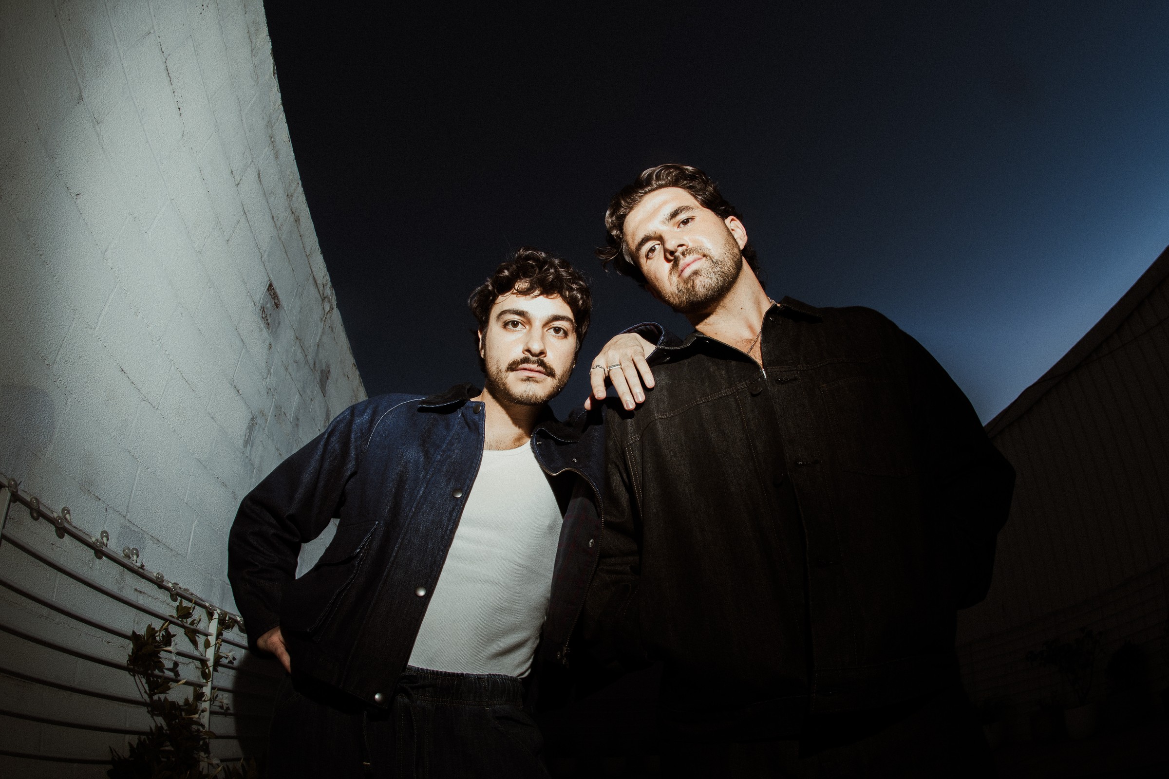 SUM SUN's Nick Benton and Ilan Pomerance, shot from a low angle against a deep blue sky beside a curved white brick wall; one wears a dark denim jacket over a white tank top, the other a black jacket with a hand on his bandmate's shoulder