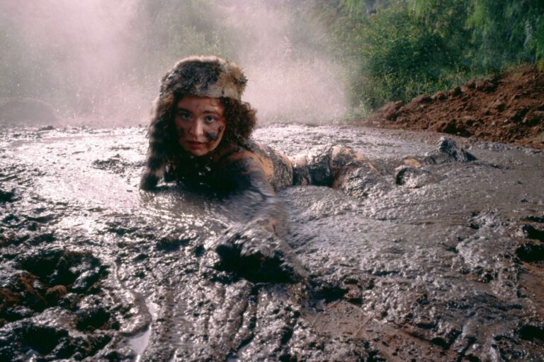 Remi Wolf press photo, lying on her stomach in a pool of wet mud wearing a fur trapper hat, her body coated in mud and mist rising behind her