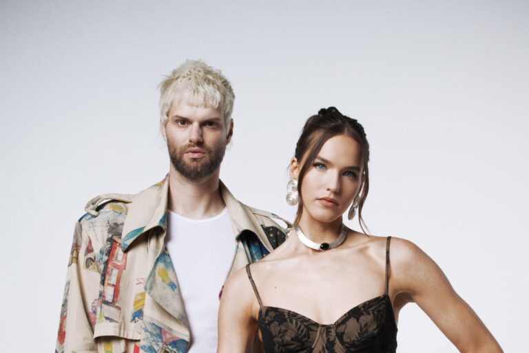 SOFI TUKKER press portrait: Tucker Halpern with bleached blond hair in a painted-print trench coat over a white tank, beside Sophie Hawley-Weld in a black lace top, silver choker and swirl earrings, against a pale grey backdrop