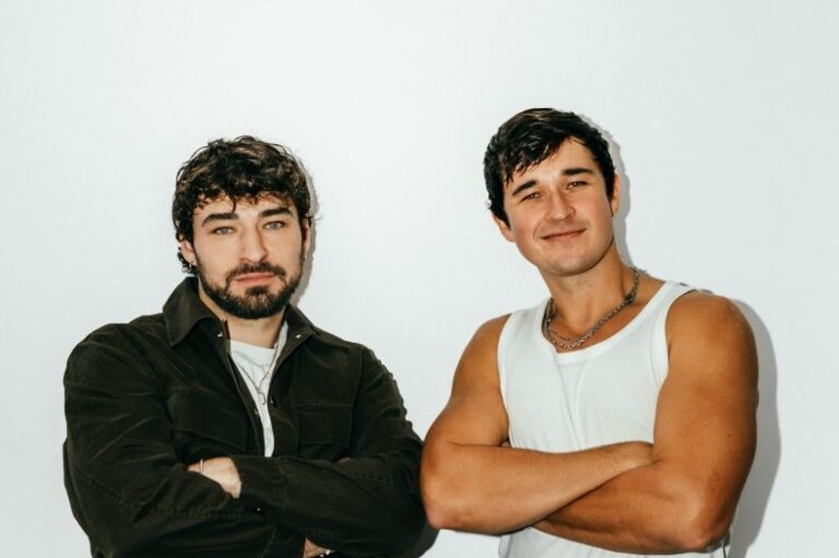 John Summit and Crankdat press photo, standing with arms crossed against a white wall, one bearded in a dark jacket over a white tee, the other in a white tank top and silver chain