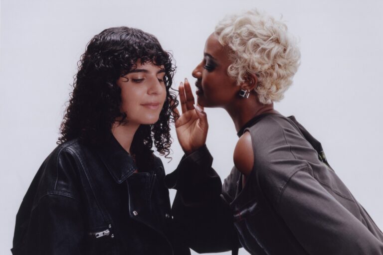 Carlita, with dark curly hair and a black leather jacket, eyes closed, as Justine Skye, with short platinum-blonde curls, leans in to whisper in her ear against a pale grey studio backdrop