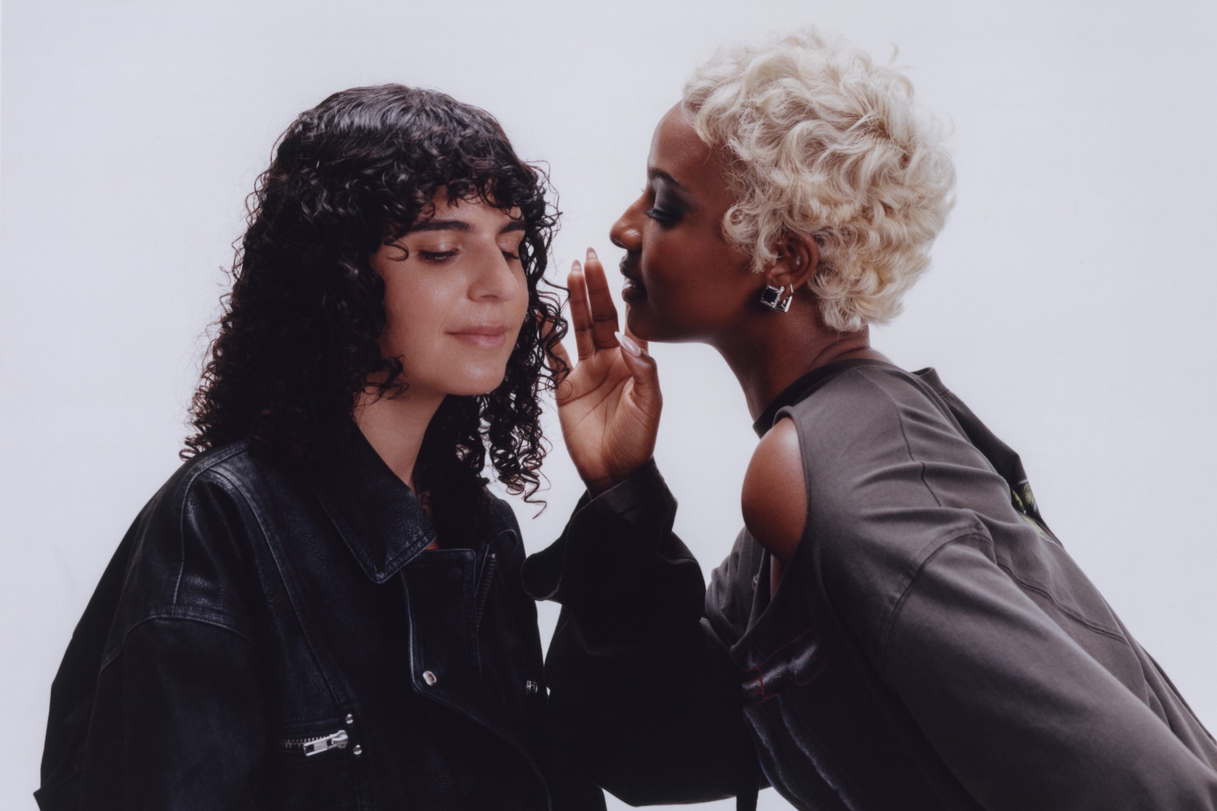 Carlita, with dark curly hair and a black leather jacket, eyes closed, as Justine Skye, with short platinum-blonde curls, leans in to whisper in her ear against a pale grey studio backdrop