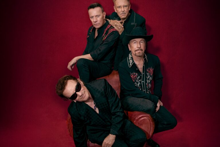 U2 press portrait of the four band members in black shirts posed around a worn red leather armchair against a deep red backdrop, Bono in front in dark sunglasses and another member in a black cowboy hat